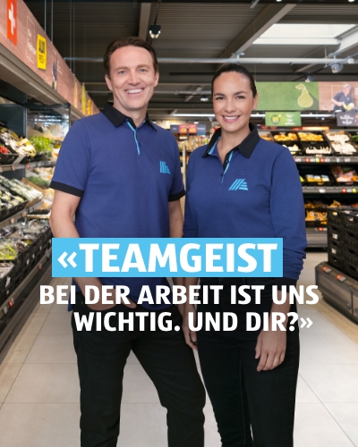 Zwei Personen ein Mann und eine Frau stehen in der ALDI Filiale nebeneinander und lächeln. Auf der Bildüberschrift steht: Teamgeist – Bei der Arbeit ist uns wichtig. Auf der Bildüberschrift steht: Teamgeist – Bei der Arbeit ist uns wichtig. Und dir?