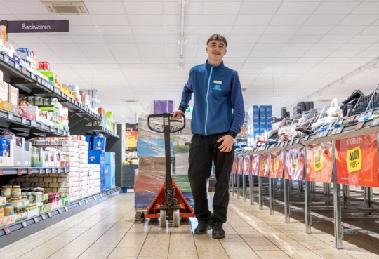 Ein Mitarbeiter rollt mit dem beladenen Palettrolli durch die Gänge einer ALDI Suisse Filiale und lächelt.