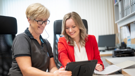Deux femmes sont assises à leur poste de travail et sourient en mangeant quelque chose devant leur plateau.
