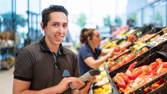 Un collaborateur d'ALDI Suisse se tient devant le rayon des légumes et contrôle l'assortiment avec une tablette.
