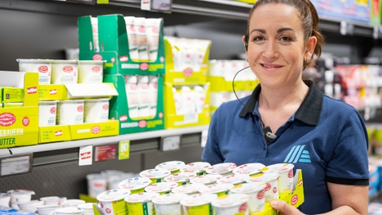 Eine Mitarbeiterin der ALDI Suisse Filiale füllt lächelnd die Regale mit Joghurt auf.