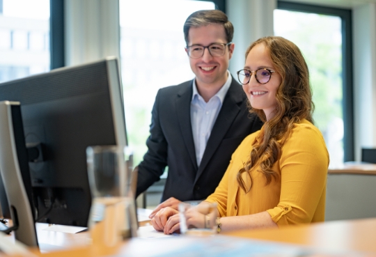Zwei Personen darunter eine Frau und ein Mann stehen am Arbeitsplatz und besprechen etwas zusammen am Desktop.