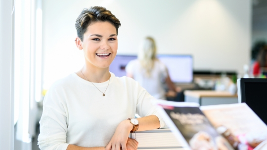 Une femme se tient à sa table de travail et sourit aimablement.