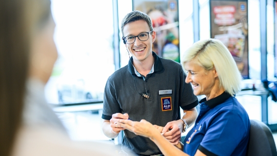Deux employés se tiennent dans une filiale d'ALDI Suisse, tous deux souriants et tenant un ticket de caisse à la main. De la troisième personne, on ne voit que l'arrière de la tête.