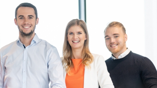 Trois personnes, dont deux hommes et une femme, se tiennent côte à côte et sourient.