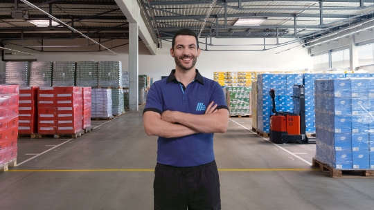Un homme se tient dans un entrepôt et sourit aimablement. Des palettes avec des produits sont visibles en arrière-plan.