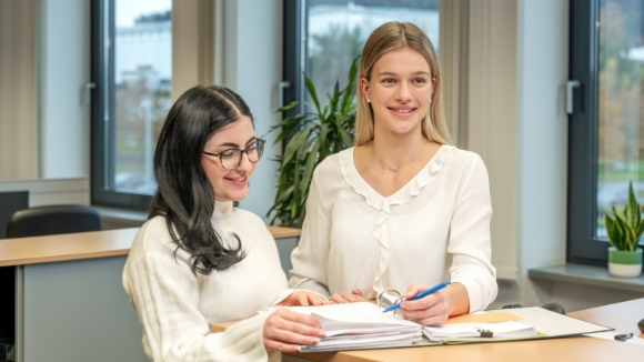 Zwei junge Frauen stehen vor ihrem Arbeitsplatz und besprechen lächelnd ein Dokument.