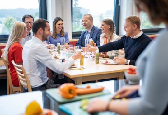 8 personnes, dont des femmes et des hommes, sont assises à une table à manger et discutent amicalement tout en mangeant des snacks sains.