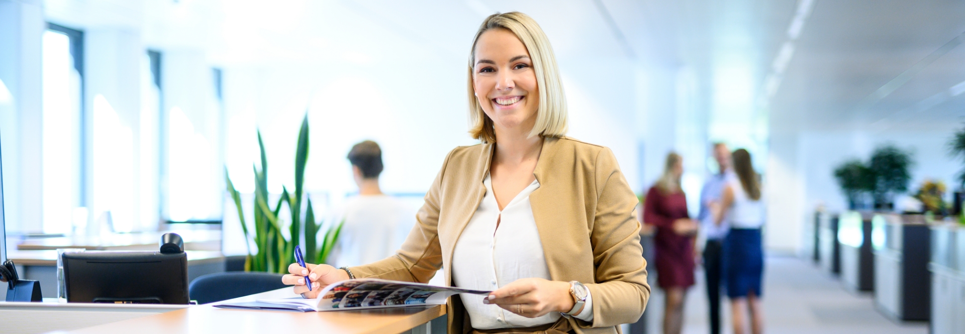 Une femme souriante se tient à un comptoir d'accueil et feuillette une brochure. En arrière-plan, on voit des personnes en train de discuter.