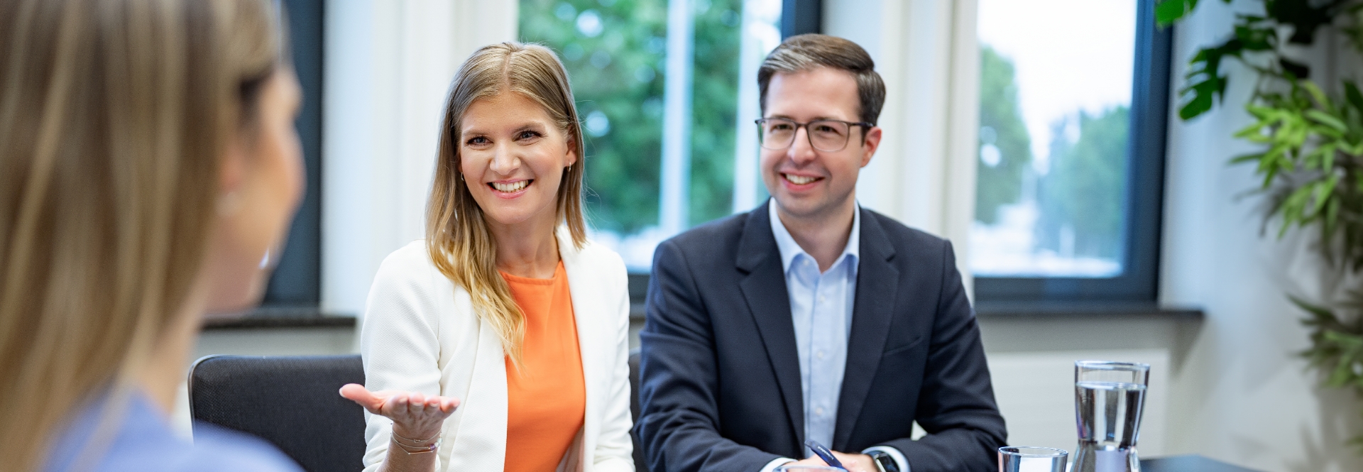 Eine Frau und ein Mann sitzen an einem Tisch und führen mit einer anderen Frau ein Bewerbungsgespräch. Beide Lächeln.