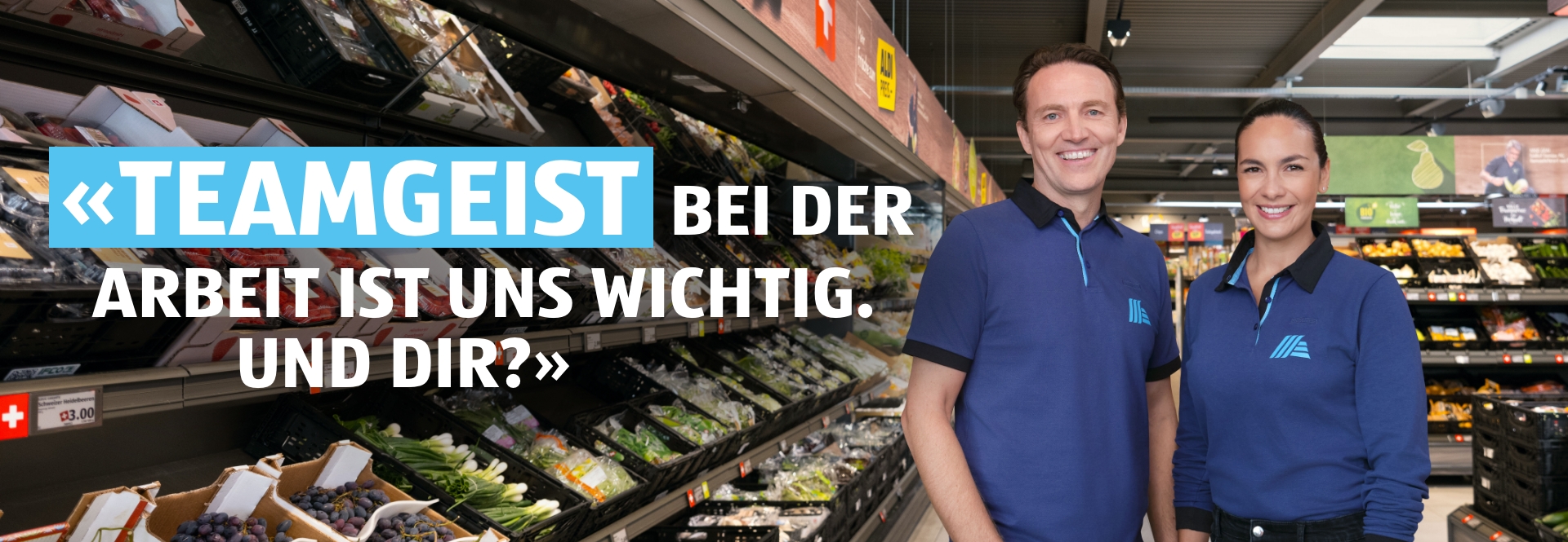 Zwei Personen ein Mann und eine Frau stehen in der ALDI Filiale nebeneinander und lächeln. Auf der Bildüberschrift steht: Teamgeist – Bei der Arbeit ist uns wichtig. Auf der Bildüberschrift steht: Teamgeist – Bei der Arbeit ist uns wichtig, und dir? 